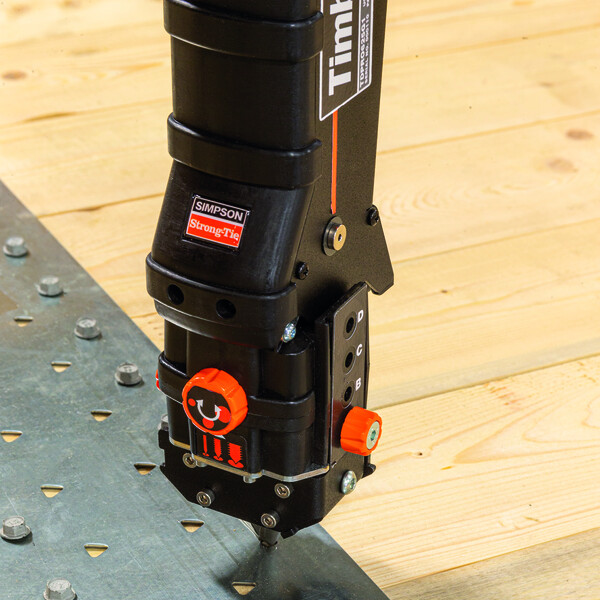 Eine Holzfläche füllt fast das ganze Bild aus, nur unten links ist ein Stück Metallband mit Löchern für Schrauben oder Nägel zu sehen. Einige der Löcher sind bereits mit Sechskantschrauben ausgefüllt. Ein Timber Drive, dessen unterer Abschnitt von oben ins Bild ragt, setzt an, mit der Präzisionsspitze eine weitere Schraube zu installieren.